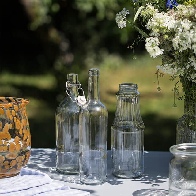 Tischdeko für Gartenparty - Step 2 Drei leere, saubere Glasflaschen stehen auf einem Tisch im Garten, bereit zum Befüllen mit Wasser und Blumen.
