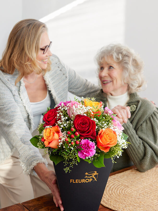 Ältere Frau freut sich über einen farbenfrohen Blumenstrauß zum 80. Geburtstag, den ihre Tochter überreicht.