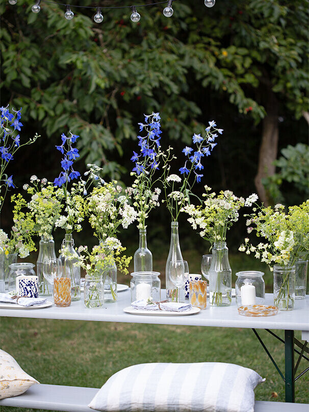 Detailaufnahme eines sommerlich gedeckten Tisches mit Wildblumen in Flaschenvasen, bunten Gläsern und Kerzen im Glas.