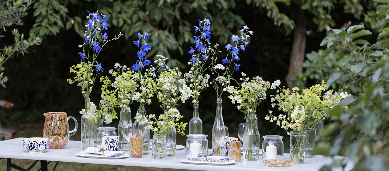 Ein sommerlich gedeckter Tisch im Garten mit vielen Glasvasen und Wildblumen, arrangiert für ein festliches Outdoor-Dinner.