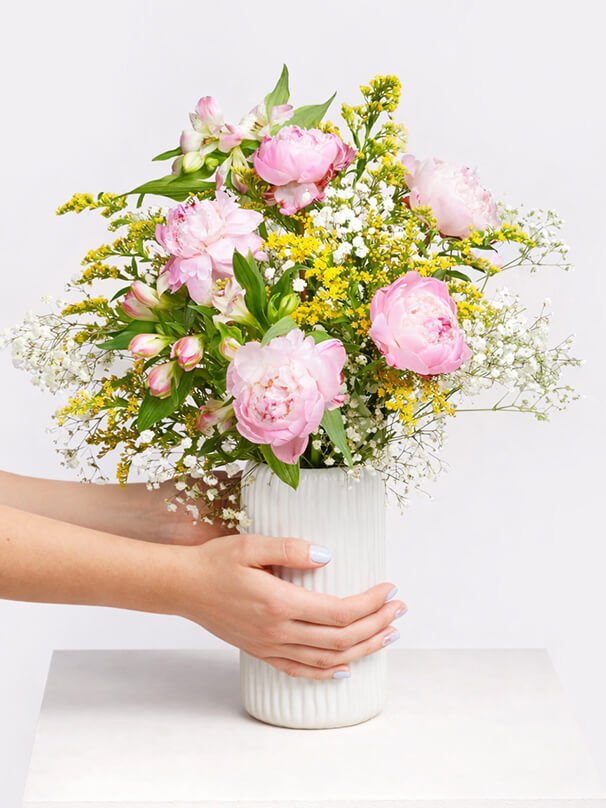Hände halten eine weiße Vase mit rosa Pfingstrosen und gelben Blüten als Geburtstagsgeschenk zum 18 Geburtstag