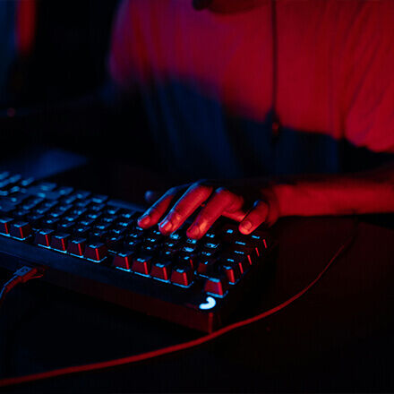 Eine Hand beleuchtet von rotem und blauem Licht liegt auf einer Tastatur, ein intensives Gaming-Setup im Dunkeln.