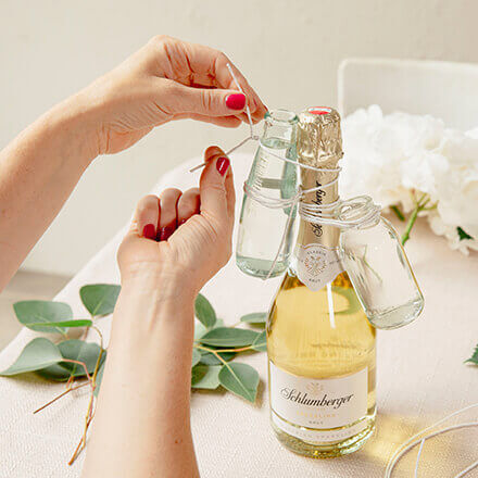 Eine Person befestigt kleine Glasvasen mit Blumendraht am Hals einer Sektflasche, die auf einem Tisch steht. Im Hintergrund sind weiße Blumen und Eukalyptuszweige zu sehen, die für die Dekoration verwendet werden sollen.
