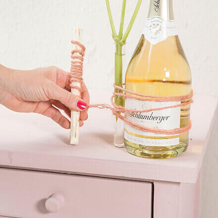 Ein Glasröhrchen wird mithilfe einer rosafarbenen Schnur an einer Schlumberger Sektflasche befestigt. In dem Glasröhrchen befindet sich Wasser und eine Dahlie ragt hinaus.