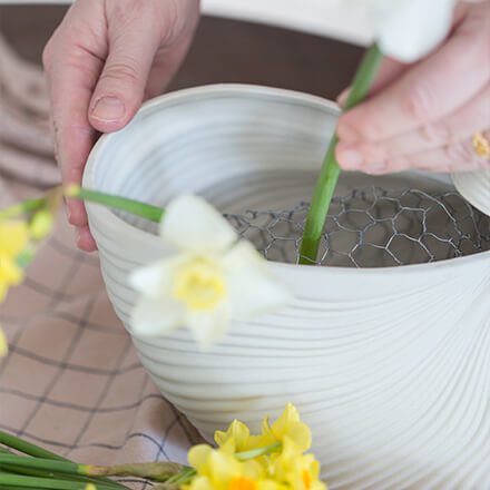 Frühlingsblumen werden in eine Steckhilfe aus Hasendraht arrangiert, welche sich in einer bauchigen Vase befindet