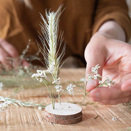 Eine Hand steckt mehrere kleine Trockenblumen in Löcher in einer kleinen, dünnen Holzscheibe