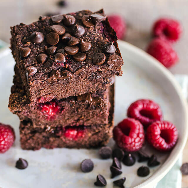Schokoladen-Brownies mit Himbeeren und Schokostückchen auf einem weißen Teller, umgeben von frischen Himbeeren