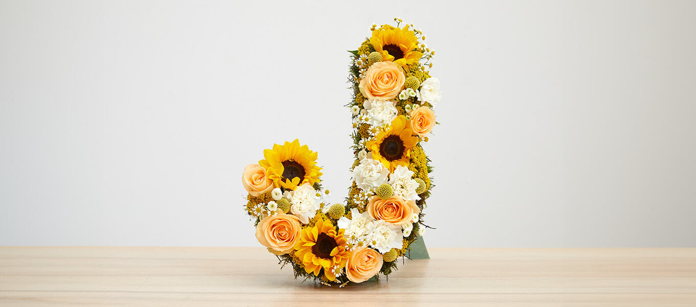 Ein dekorativer Buchstabe "J" aus frischen Blumen, darunter Sonnenblumen, Rosen und andere Blüten, auf einem Holztisch. Die Blumen sind dicht aneinander gereiht und bilden eine lebendige und farbenfrohe Gestaltung.